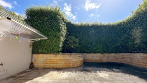 A paved courtyard with a brick wall, a white building, and a dense hedge.