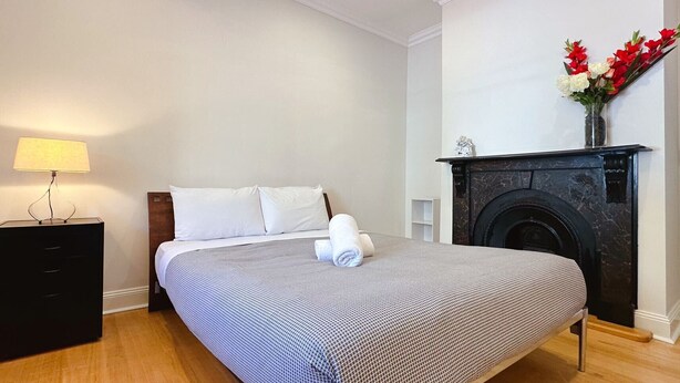 A hotel room with a bed, a nightstand, a lamp, a fireplace, and a vase with flowers.