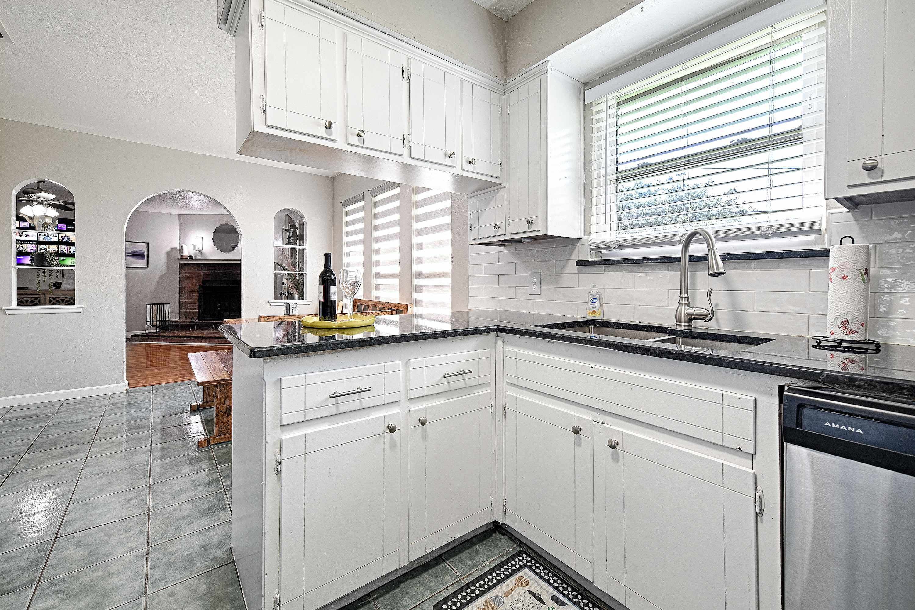 A kitchen with white cabinets, a black countertop, a dishwasher, and a window with blinds.