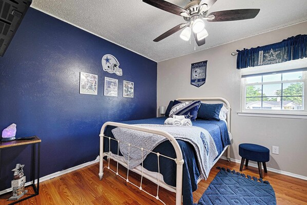 Une chambre à coucher comprenant un lit, un ventilateur de plafond, une fenêtre avec des rideaux et des éléments de décoration murale, notamment le logo des Dallas Cowboys.