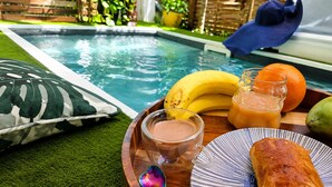A poolside setting with a wooden tray containing a baguette, bananas, a glass of orange juice, and a smoothie.