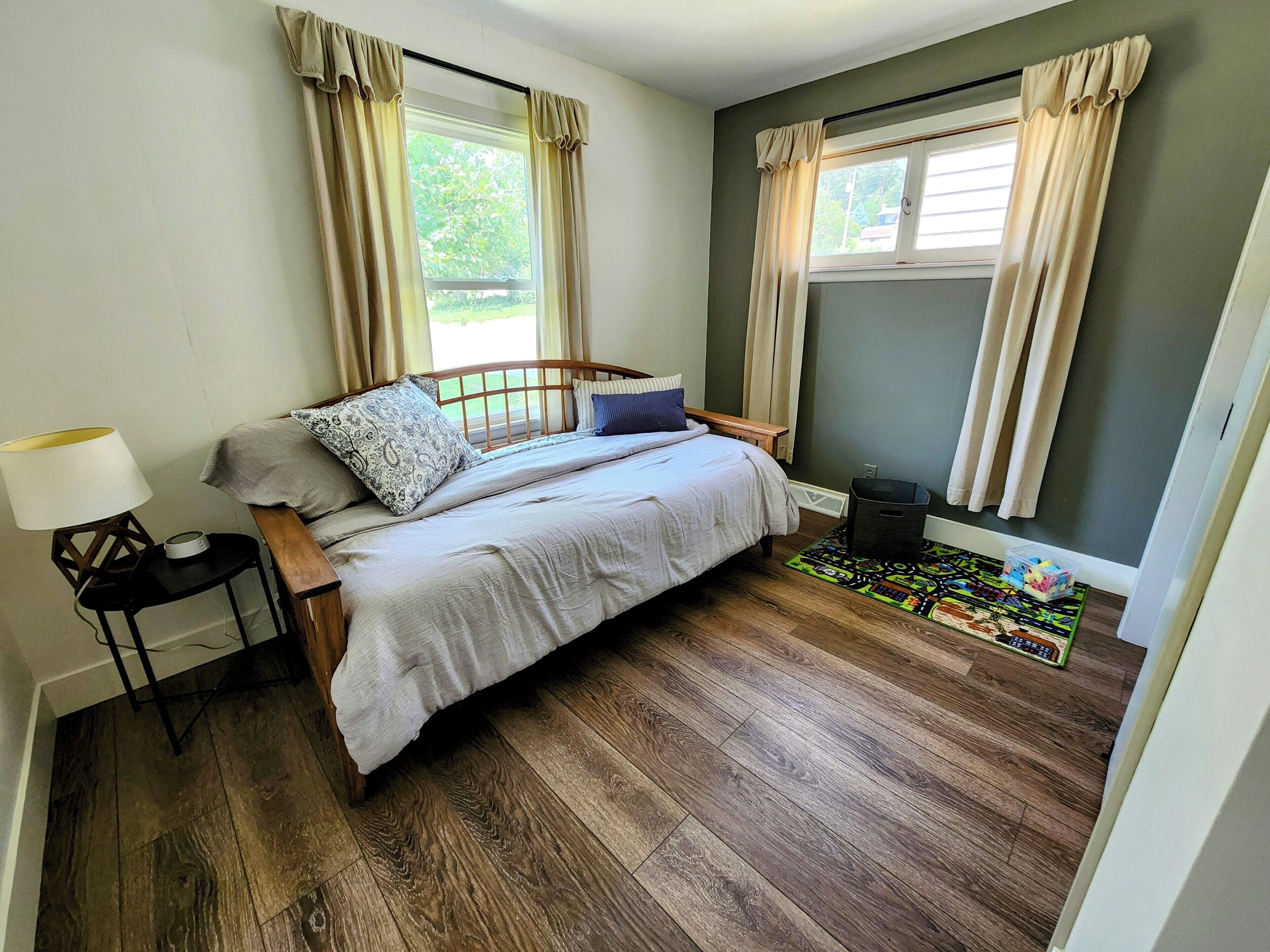 A bedroom with a wooden bed, a nightstand, a lamp, a window with curtains, and a small rug.