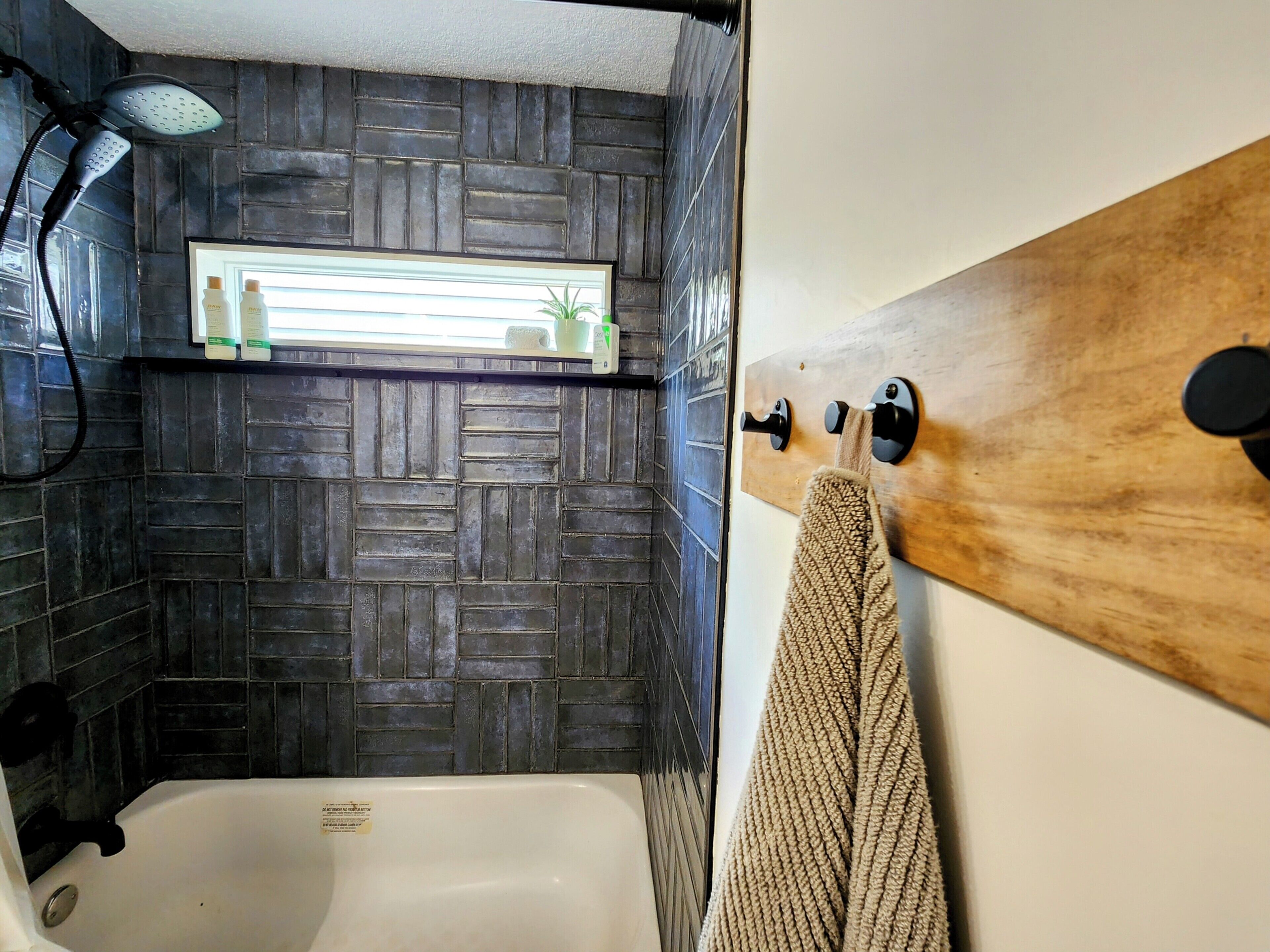 Un baño con bañera, ducha, paredes de azulejos oscuros, un estante de madera con artículos de aseo y una toalla colgada de un gancho.