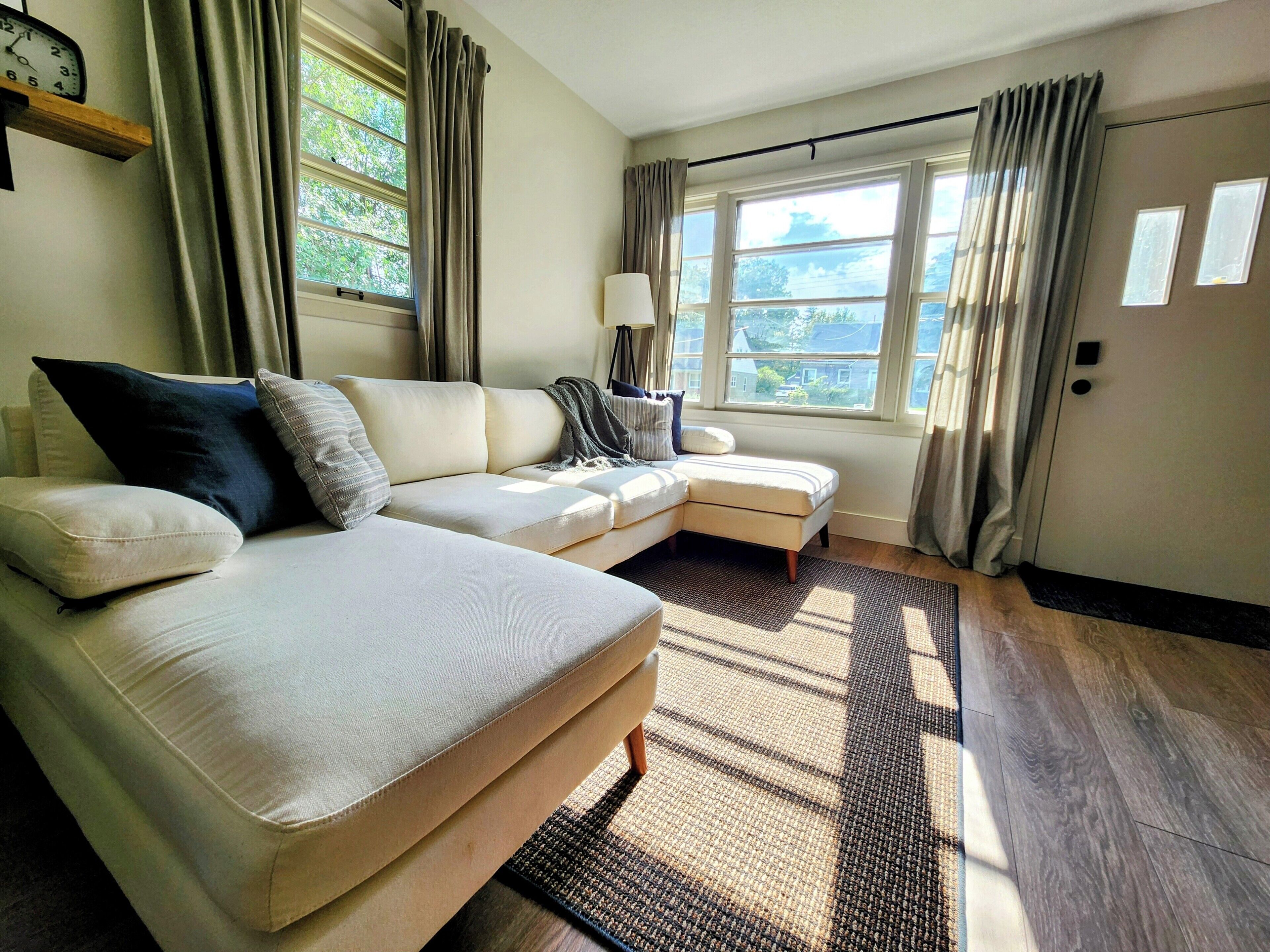 A modern living room with a sectional sofa, a small ottoman, a floor lamp, and large windows with curtains.