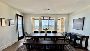 A spacious dining room with a large wooden table, multiple chairs, and a view of the outdoors through large windows. - Newly Built Gem with Panoramic Lake Views and Chic Interiors (Garden City)