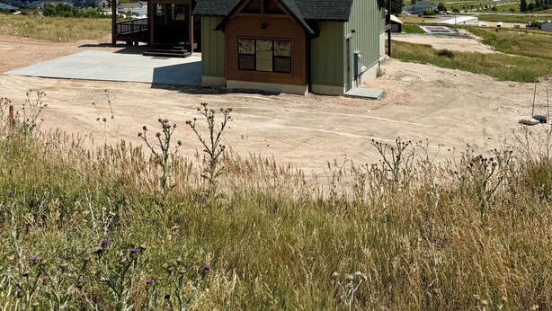 A two-story house with a green roof and a small porch, situated on a dirt driveway with a grassy field in the foreground and a body of water in the background. - Newly Built Gem with Panoramic Lake Views and Chic Interiors (Garden City)