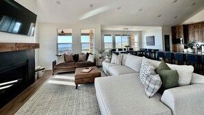 A spacious living room with a fireplace, a large sofa set, and a dining area with a view. - Newly Built Gem with Panoramic Lake Views and Chic Interiors (Garden City)