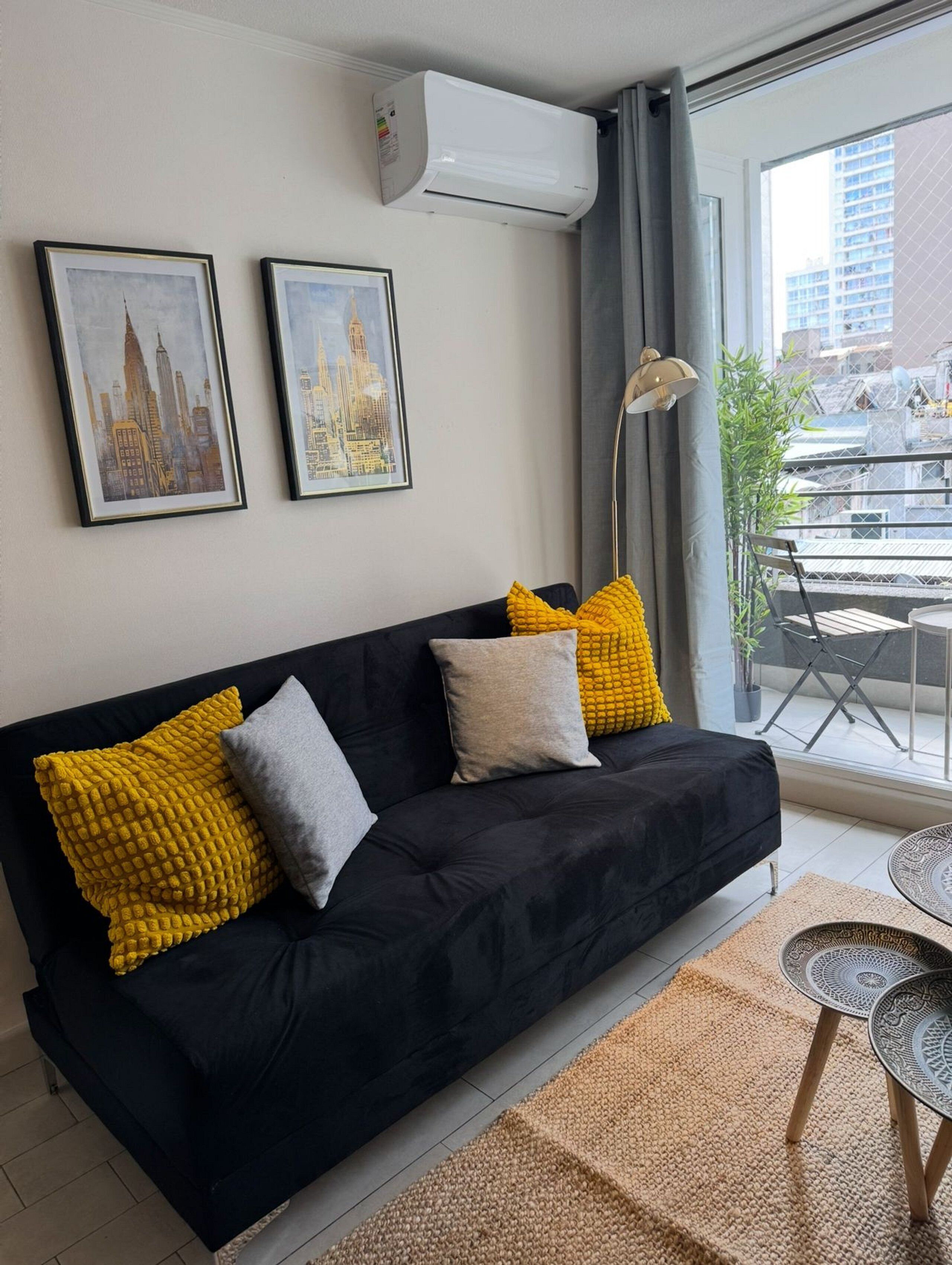 Un salon moderne avec un canapé noir, deux coussins jaunes à texture et un coussin gris. On peut voir deux tableaux représentant des paysages urbains, encadrés, accrochés au mur, ainsi qu’un climatiseur blanc et une lampe sur pied. Une petite table ronde recouverte d’une nappe à motifs est visible au premier plan.