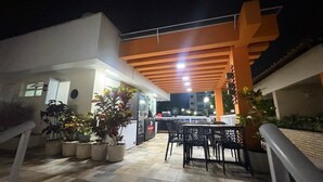 An outdoor patio area with a dining table and chairs, potted plants, and a building with a modern design. - Barra Olímpica Riocentro Apartment (Camorim Rio de Janeiro)