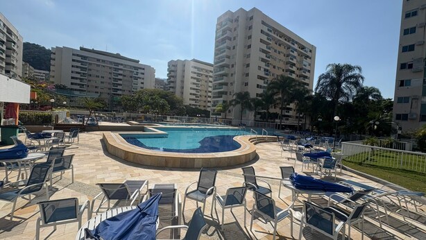A swimming pool with a curved design, surrounded by lounge chairs and umbrellas, with tall residential buildings in the background. - Barra Olímpica Riocentro Apartment (Camorim Rio de Janeiro)