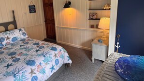 A bedroom with a bed, a nightstand, a lamp, a shelf with decorative items, and a door. - Escape to this charming beach cottage along the coast of Broadkill Beach (Milton)