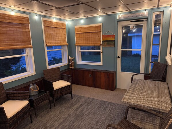 A cozy room with wicker furniture, a small table, and a view of the outdoors through large windows with blinds.
