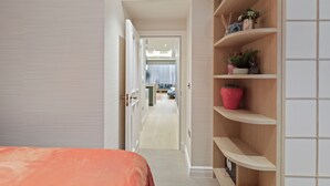 Un pasillo de hotel con una cama a la izquierda, un mueble de madera con objetos decorativos a la derecha y una vista parcial de un baño en la distancia.
