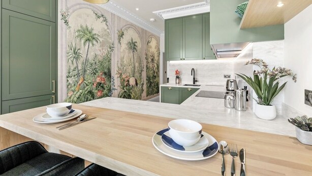 Cocina moderna con armarios verdes, encimera de mármol y comedor con una mesa de madera para dos.