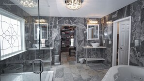 A modern bathroom with marble walls, a glass shower enclosure, a large bathtub, and a walk-in closet with shelves.
