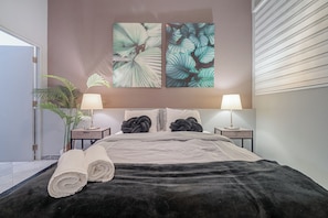 Une chambre à coucher moderne avec un grand lit, deux lampes de chevet, une plante en pot et des œuvres d’art accrochées au mur.