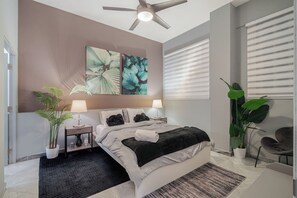 Un dormitorio moderno con una cama, dos mesitas de noche con lámparas, un ventilador de techo y ventanas amplias con persianas.