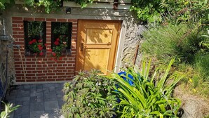 A brick building with a wooden door, two windows with red flowers, and a blue container.