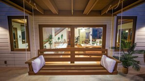 A wooden bench with white cushions in a well-lit outdoor area with a view of an interior space.