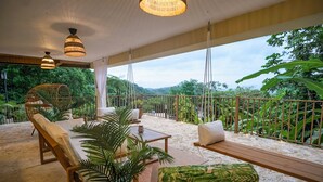 A covered outdoor seating area with wooden furniture and hanging lamps, overlooking a lush landscape.