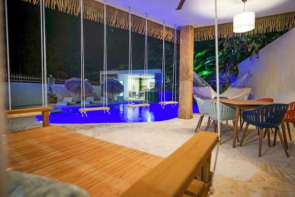 A poolside area with a wooden table and chairs, a hammock, and a view of a building through large windows.