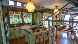 A spacious kitchen with wooden countertops, a central island, and bar stools. There are hanging lights and a view of greenery outside.