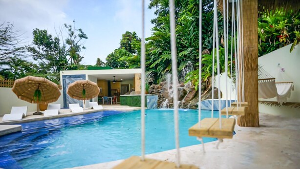 A swimming pool with lounge chairs and umbrellas, surrounded by tropical plants and a waterfall feature.