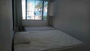 A small, dimly lit room with two single beds, a window with a view of a building and palm trees, and a white cabinet.