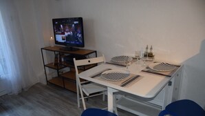 A dining area with a white table, blue chairs, a television, and a shelf with books and a lamp. - L' Escale, appartement climatisé à 50 mètres de la plage avec WIFI et Box Tv (Fos-sur-Mer)