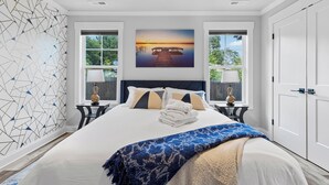 Une chambre moderne avec un grand lit, deux tables de chevet, une tête de lit et une vue sur un coucher de soleil sur l’eau.