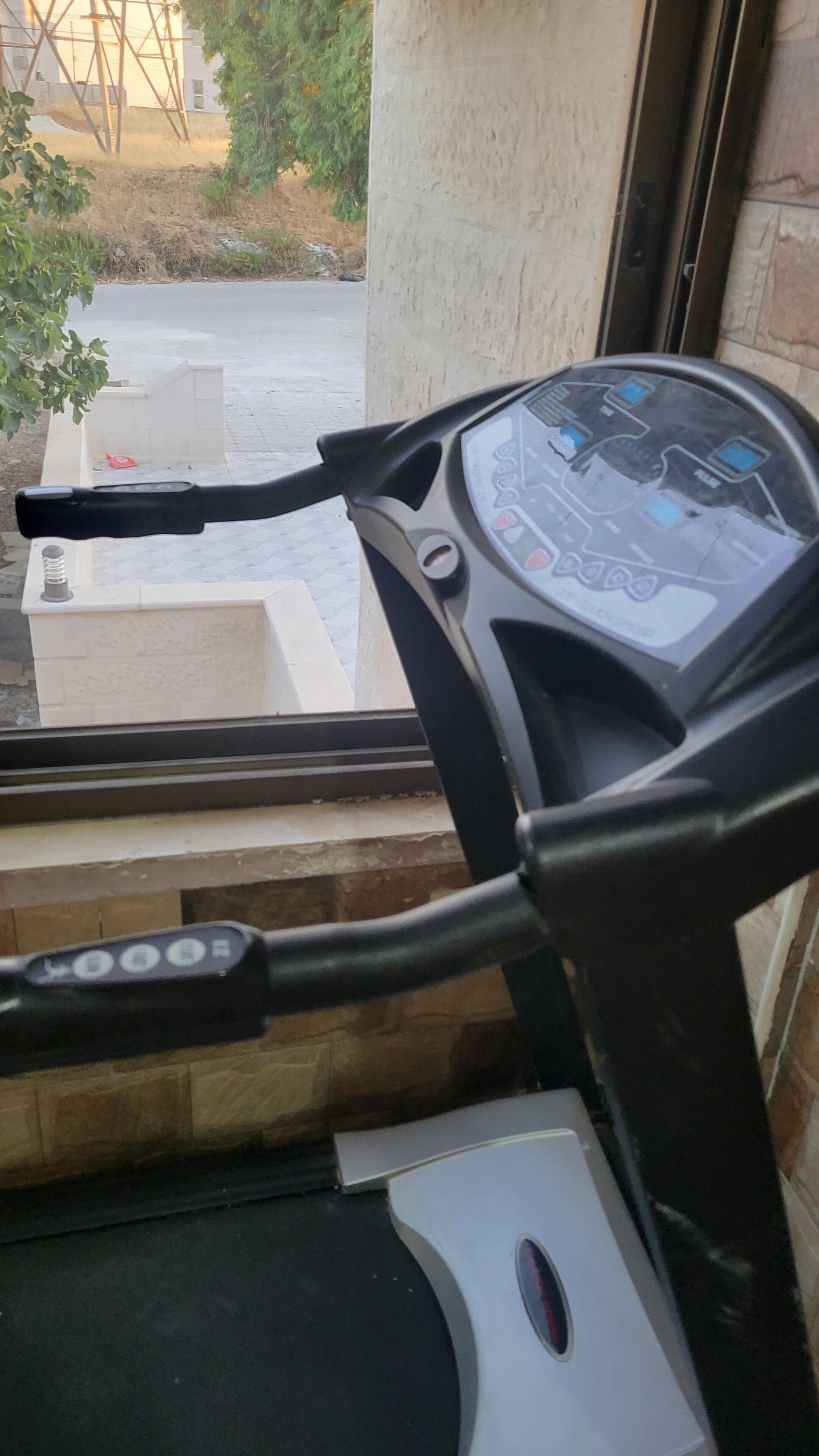 A treadmill with a control panel, a window with a view of a building and greenery outside.