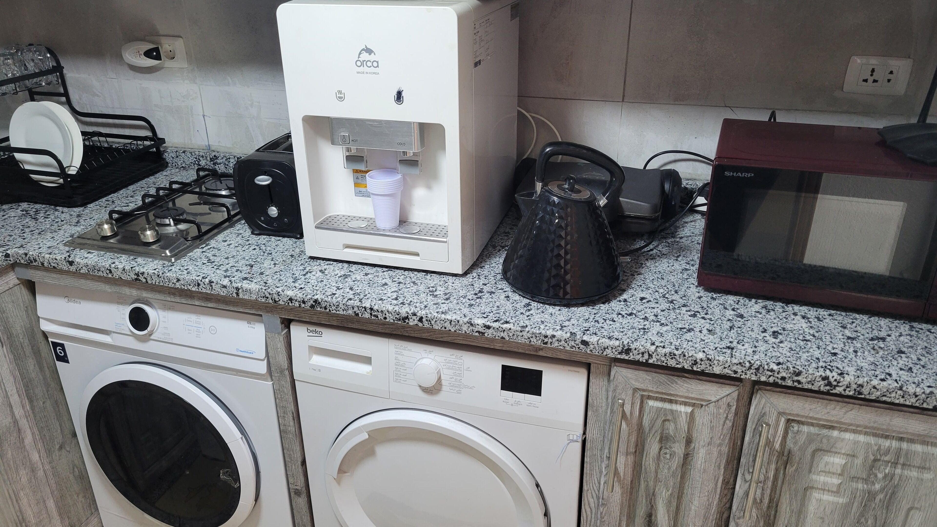 A kitchen with a washing machine, a coffee maker, a microwave, and a dish rack.