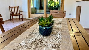 Una mesa de madera con mantel decorado, una planta en maceta y un banco en una habitación con puerta corredera de vidrio que da a un espacio exterior.