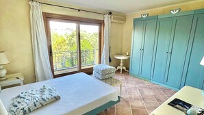 Un dormitorio con cama, una mesita, una silla, un armario y un balcón con cortinas.