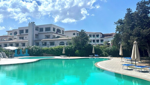 Una piscina con sillones de descanso y sombrillas, rodeada por un edificio con varios balcones y un cielo azul despejado.