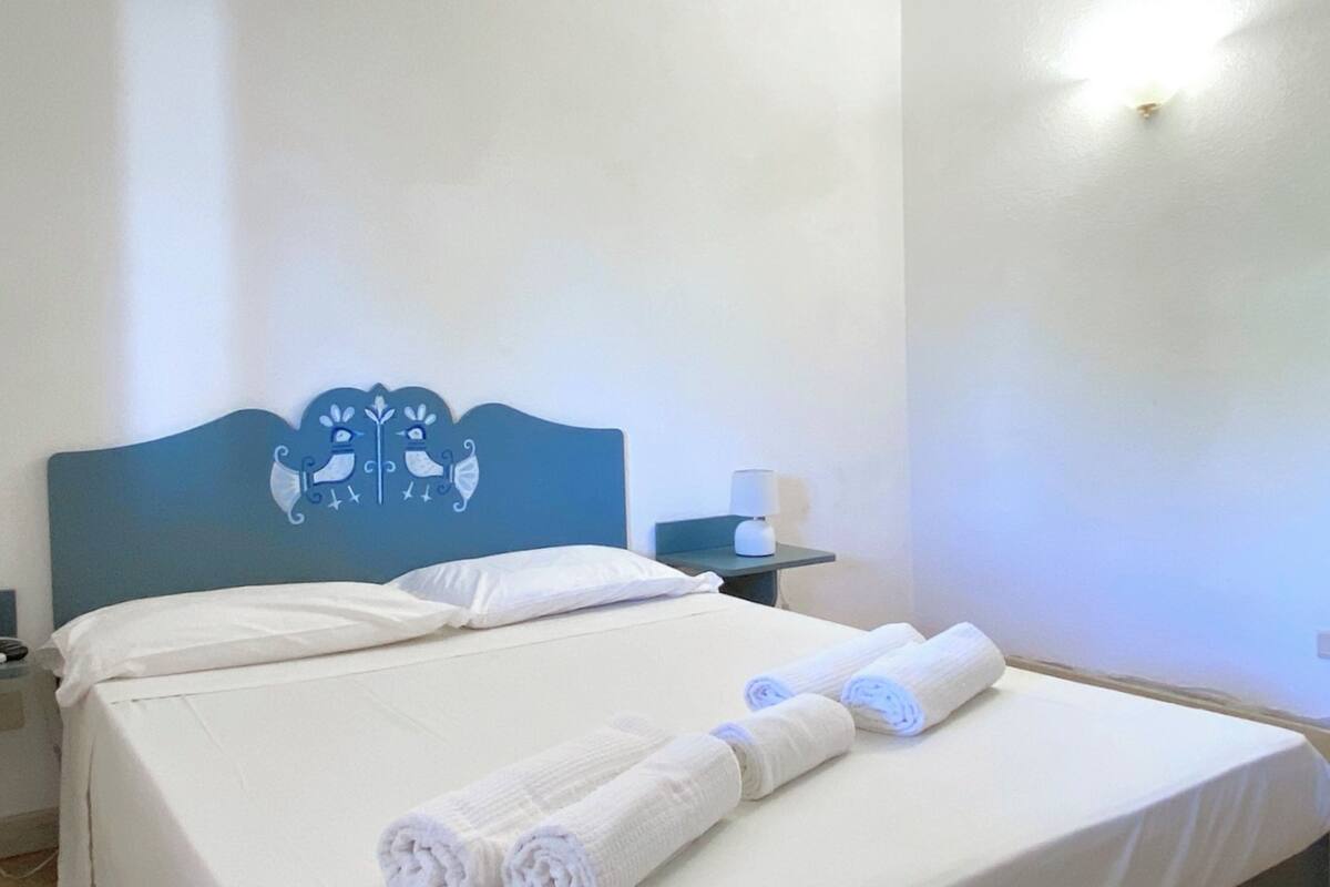 Una cama bien hecha con sábanas blancas y toallas enrolladas, un cabecero azul con un diseño de pájaro, una mesita de noche con una lámpara y una lámpara de pared.