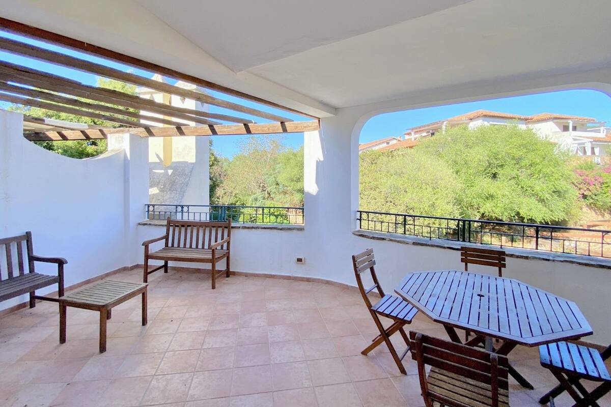 Un patio techado al aire libre con mesa y sillas de madera, un banco de madera y vistas a edificios y árboles.