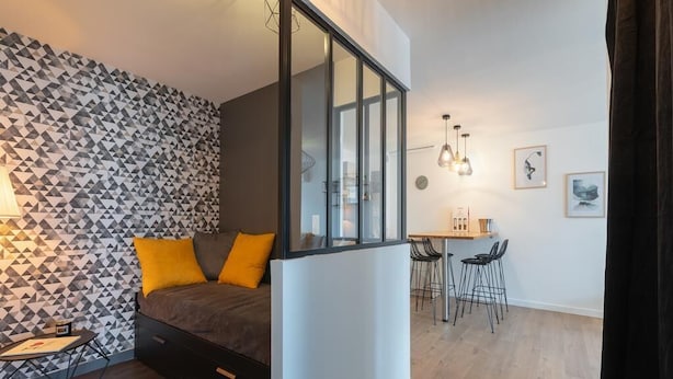 A modern bedroom with a geometric-patterned wall, a bed with yellow pillows, a desk with two chairs, and a dining area with a small table and chairs.