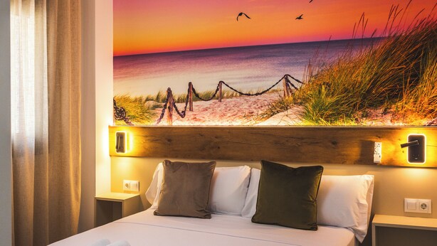 Een hotelkamer met een bed, kussens, een nachtkastje en een muurschildering van een strand.