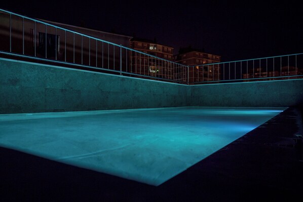 Zona de piscina al aire libre de noche, con barandilla metálica y vista a edificios al fondo.