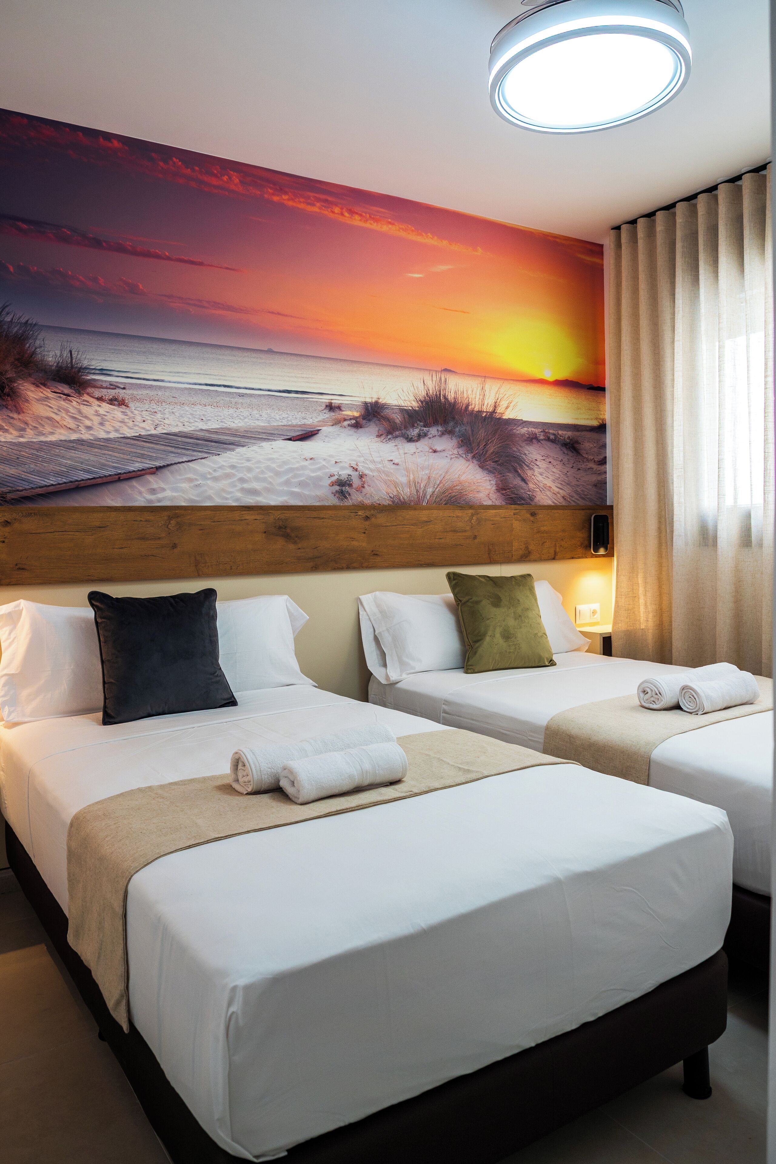 A hotel room with two beds, a wooden headboard, a sunset mural, and a window with curtains.