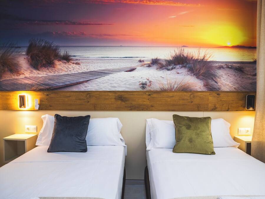 Habitación de hotel con dos camas, cada una con almohada y mesita de noche. Un cuadro grande de una escena de playa está sobre las camas.