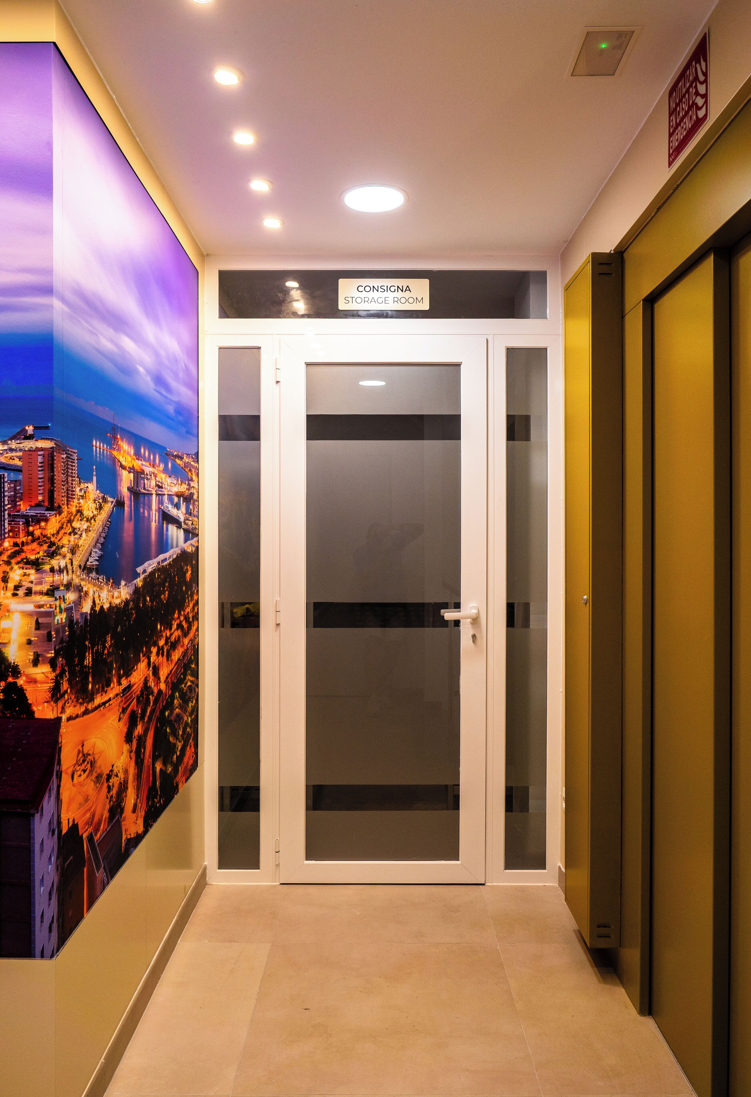 Ein Hotelkorridor mit einem Schild mit der Aufschrift 'Consigna Storage Room', einer Glastür und Blick auf eine nächtliche Stadtansicht.