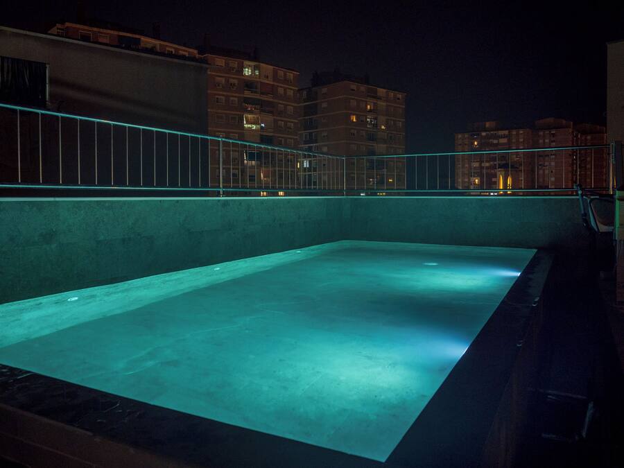 A rooftop swimming pool with a glass barrier and a view of surrounding buildings at night.