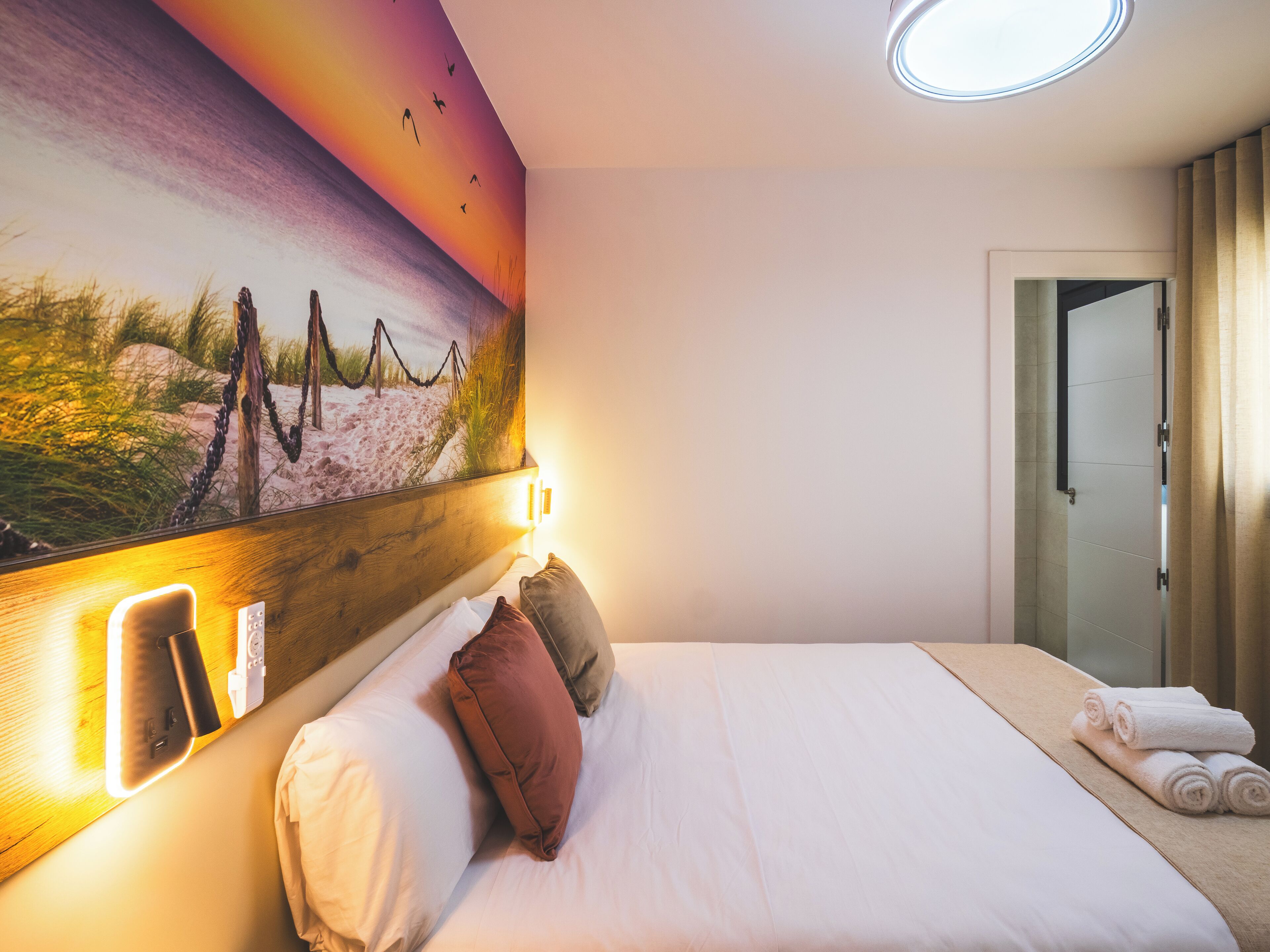 A hotel room with a bed, pillows, a painting of a beach scene, a bedside table with a phone and charger, and a door leading to a bathroom.