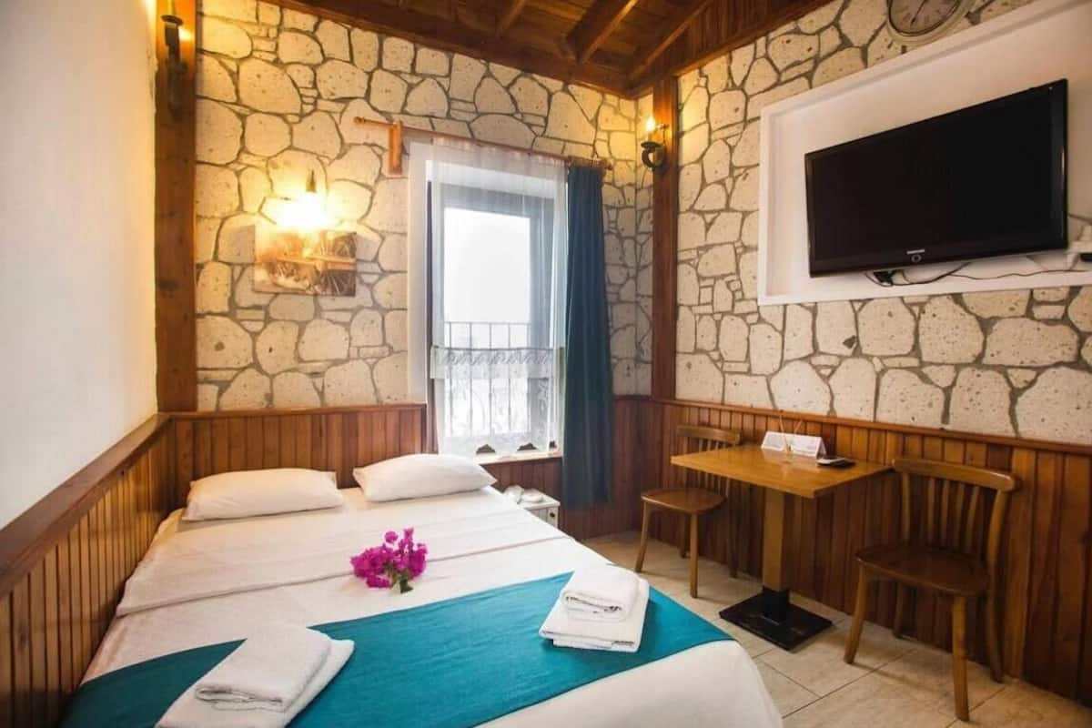 Una habitación de hotel con pared de piedra, cama con colcha color turquesa, escritorio de madera y televisor montado en la pared.