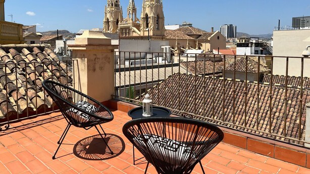 A rooftop terrace with tiled flooring, two black wire chairs, a small round table, and a view of a historic building with three spires.