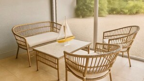 A wicker furniture set with a glass table and chairs, a small sailboat model, and a view of greenery through a window.