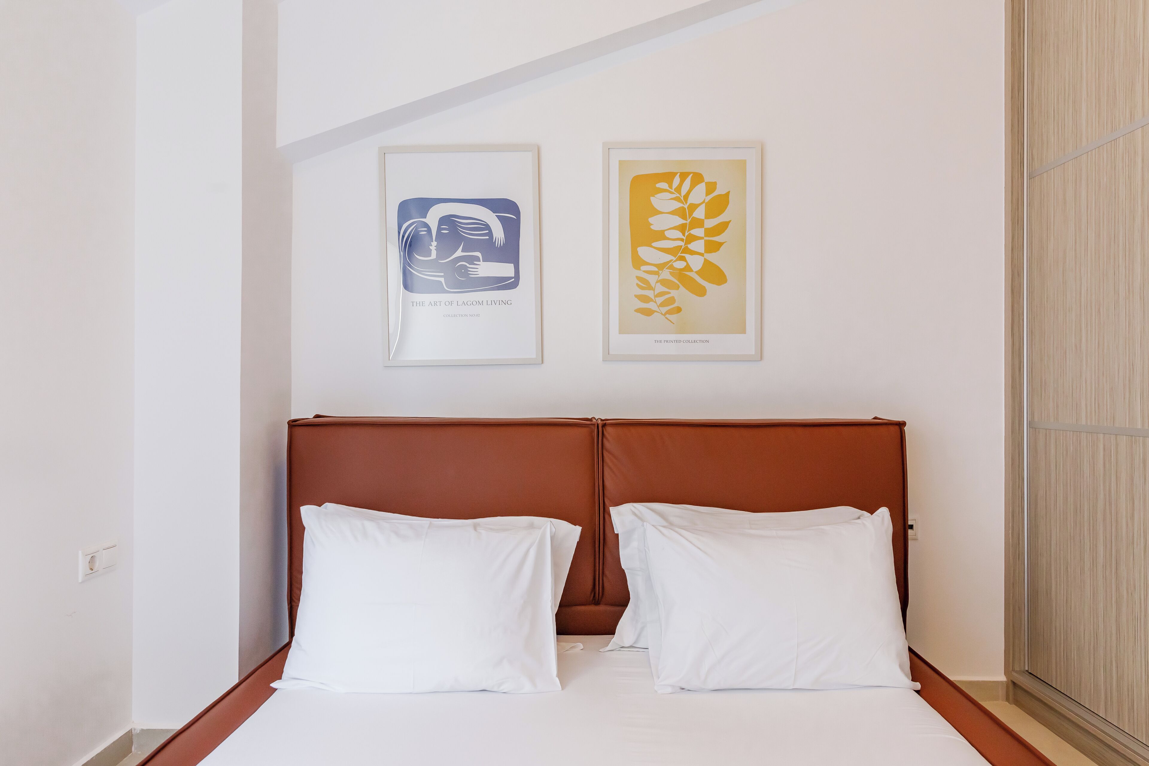 Ein ordentlich bezogenes Bett mit braunem Kopfteil und weißen Kissen in einem Zimmer mit weißen Wänden. Zwei gerahmte Kunstwerke hängen über dem Bett.