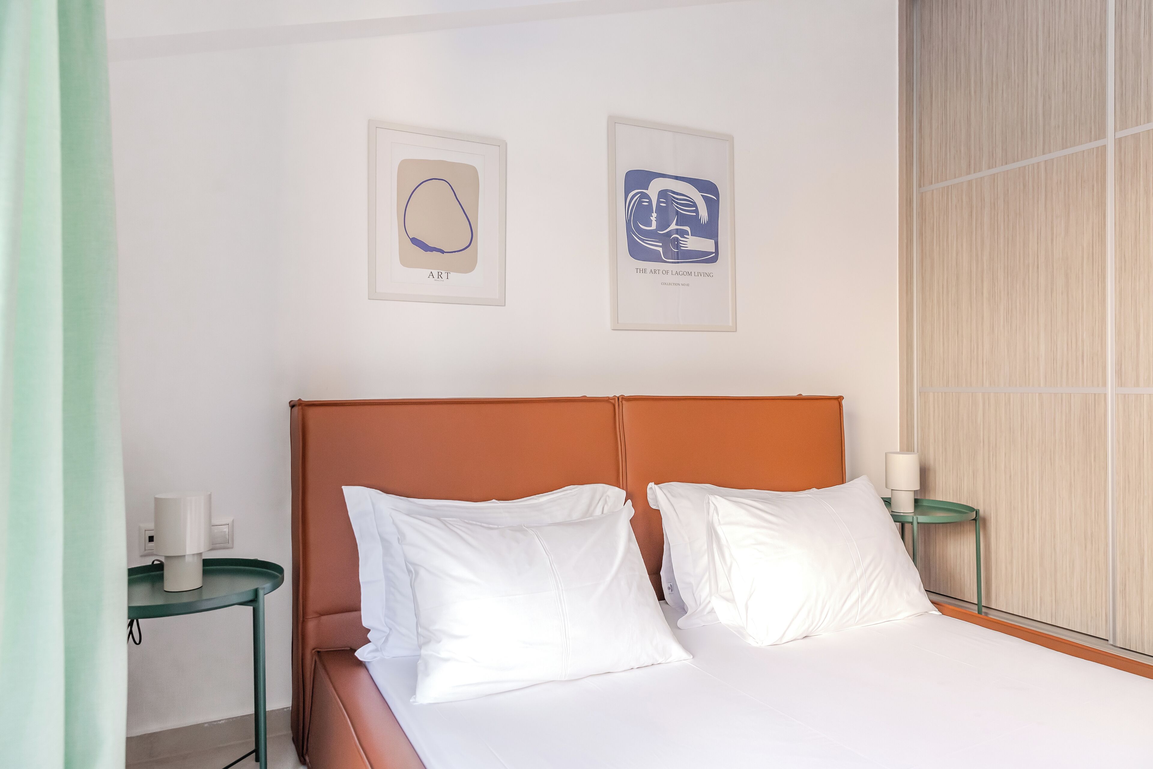 Une chambre d’hôtel avec un lit, des oreillers blancs, une tête de lit marron, deux œuvres d’art encadrées accrochées au mur et une table de chevet verte avec une lampe.